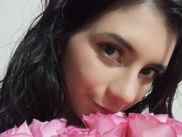 JeneKurz with dark wavy hair, brown eyes, and a subtle smile, holding pink roses, showing a cute and sexy face. Soft skin, slight blush, and a gentle expression.