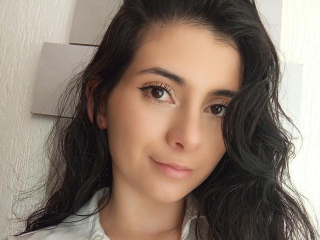 JeneKurz has tan skin, brown eyes, long wavy black hair, and a small smile. She's wearing a white shirt with a silver necklace.