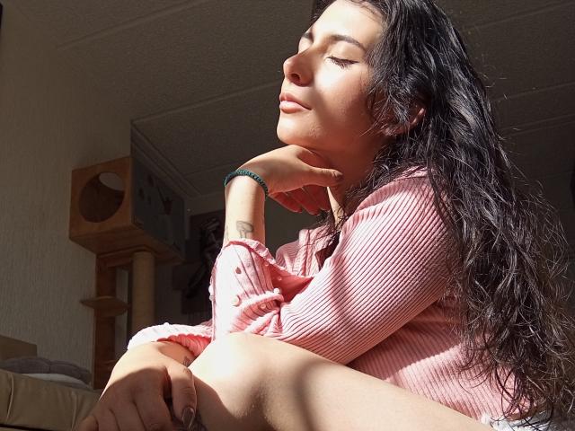 JeneKurz with long black wavy hair, eyes closed, sunlit, wearing a pink ribbed shirt, hand under chin, bracelet, tattoo on wrist, sitting with knees up, relaxed.