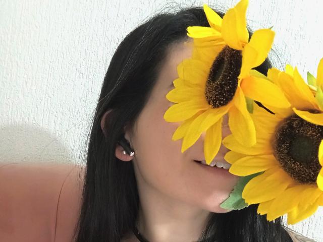 JeneKurz with black hair, face partially covered by two bright yellow sunflowers, showing a slight smile, wearing black earbuds.