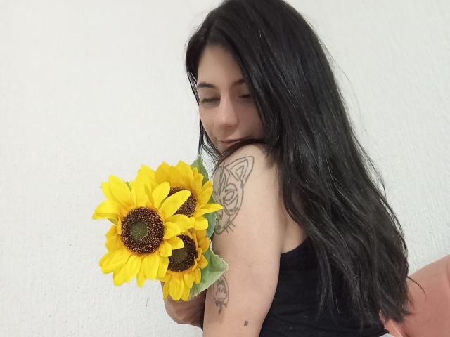 JeneKurz, met lang zwart haar, die zonnebloemen vasthoudt, heeft een tatoeage van een vlinder op haar rechterarm, draagt een zwarte tanktop, kijkt naar beneden, voor een witte muur..