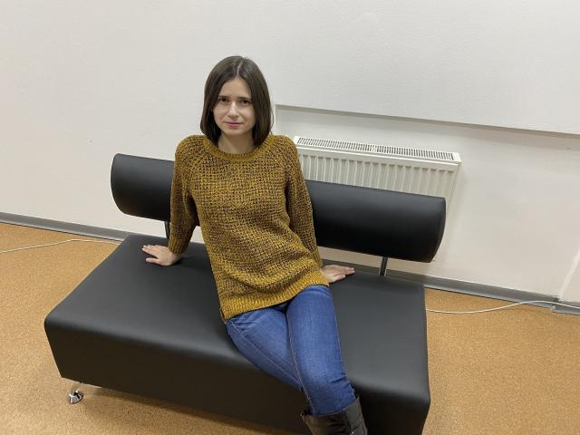 MartaLeee with brown bob, wearing mustard sweater, blue jeans, black boots, sitting on black leather bench, arms on bench, legs crossed, white wall, radiator, and carpeted floor.