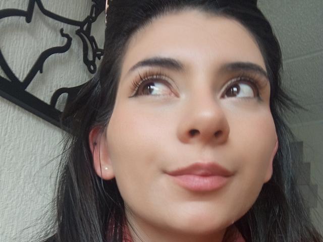 JeneKurz's face, with dark hair, brown eyes, and a slight smile, looking up with a soft expression, wearing a red shirt, and small pearl earrings.