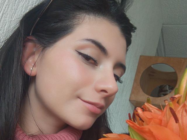 JeneKurz has smooth, pale skin, dark eyebrows, and long black hair. She's wearing a pink sweater, with orange flowers in the foreground, and a subtle smile.