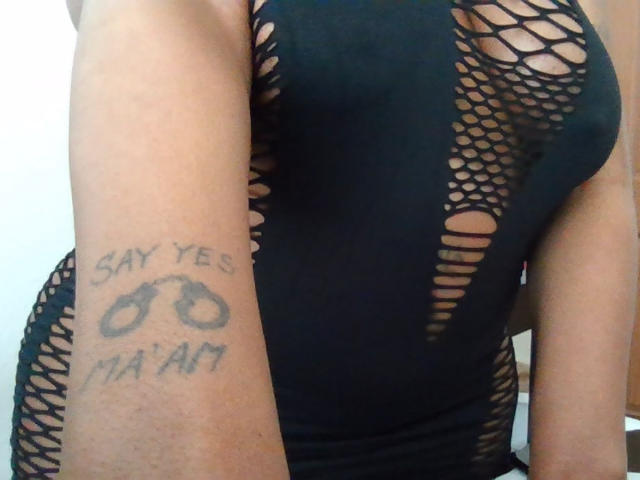 SayYesMaam's arm with a tattoo that says "Say Yes Ma'am" and handcuffs, wearing a black fishnet dress, exposing her tits and showing her brown skin.