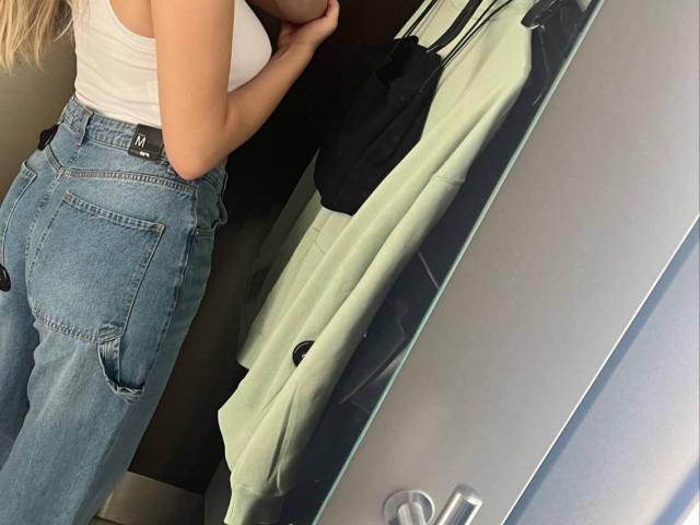 Destinis with blonde hair, white tank top, and high-waisted blue jeans, showing her ass while holding a black bag next to a green coat in a mirror reflection.