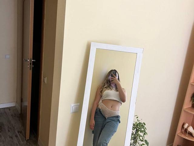 Destinis, selfie allo specchio, indossa un top corto bianco, jeans blu e biancheria intima in pizzo, mostrando il seno e i glutei, capelli biondi, tiene in mano il telefono, riflesso in uno specchio con cornice bianca, stanza..