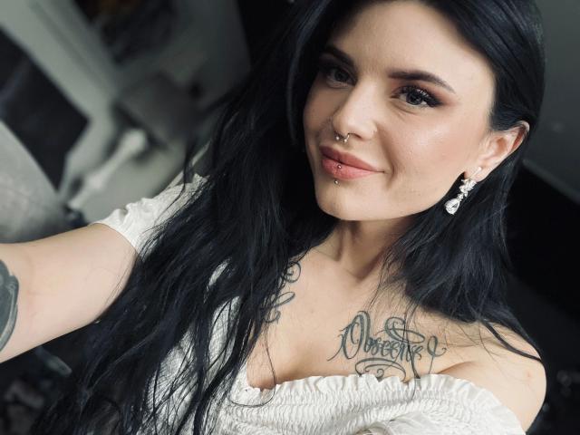 Coco1996: aantrekkelijk met zwart haar, neusring, lip piercing, oorbellen, witte top, tatoeages op de borst, lachend, telefoon omhoog houdend voor een selfie, haar borsten laten zien..
