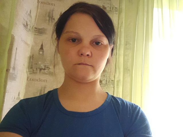 NettyEvangelina with a serious face, wearing a blue shirt, has small tits, light skin, and brown hair in a messy bun. She's standing in front of a green curtain.