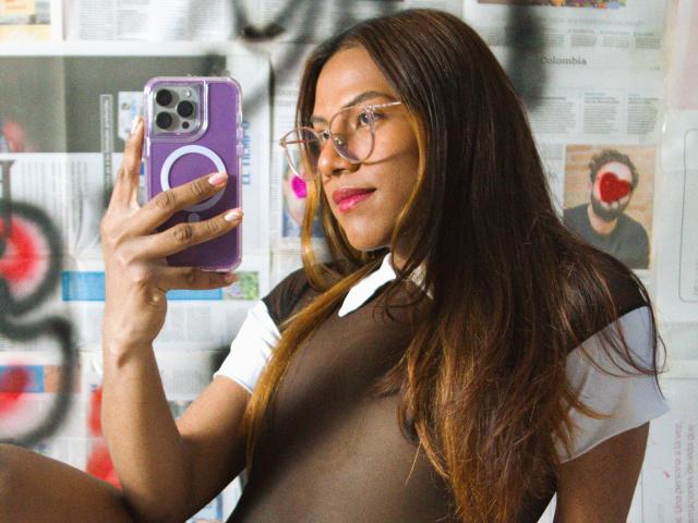 KimmySolange with long brown hair, glasses, and pink lipstick, taking a selfie with a purple phone, wearing a sheer brown top over a white shirt, showing her tits.