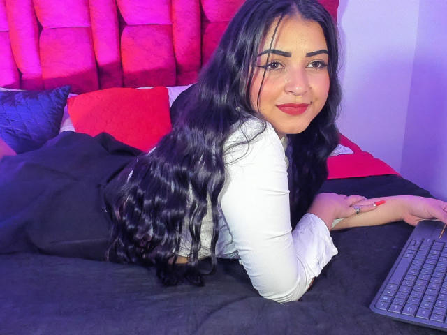 ZoeyGilmore, avec de longs cheveux noirs bouclés, une chemise blanche, du rouge à lèvres, allongée sur un lit, un pantalon noir, un clavier noir, des oreillers rouges et noirs, souriante, un anneau au nez, une bague à la main droite..