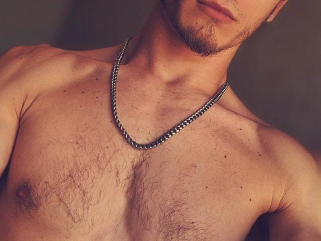 Matheo69's topless chest with a chain necklace, showing hairy skin, small nipples, and a partial beard, in a warm, brown-tinted photo. Matheo69's face is partially visible.