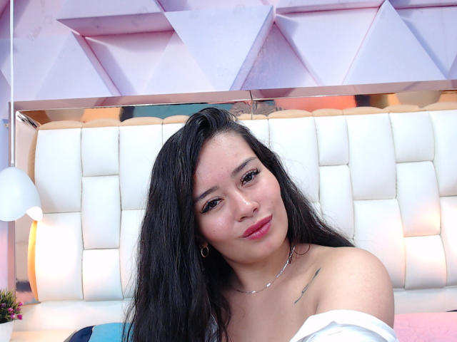 MelissaCaty with long black hair, red lipstick, small hoop earrings, necklace, and off-shoulder white top, showing a small tattoo on her right shoulder.