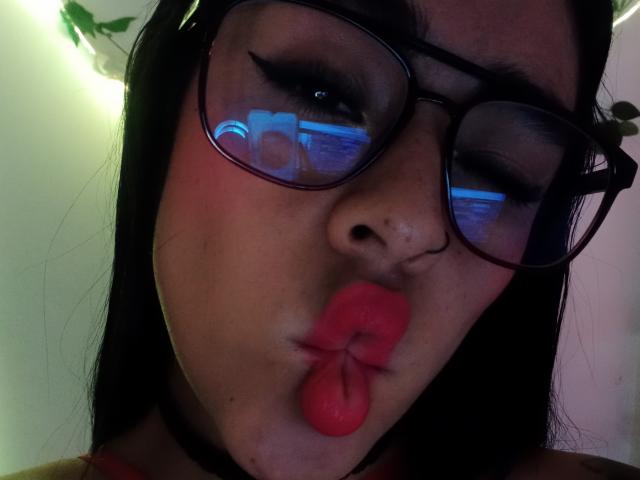 StellaBeckker with pouty red lips, black choker, and glasses, winking. Reflection in glasses shows blue light. Her face is centered, with long black hair.