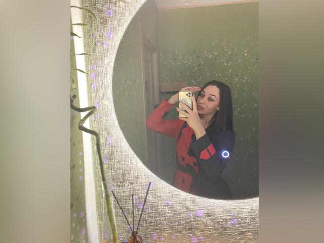 MissXPinkX mit schwarzen Haaren, in einem roten und schwarzen Kleid, hält ein Handy und macht ein Selfie im Spiegel, wobei ihre Brüste und ihr Hintern zu sehen sind. Grüne Wand mit weißen Flecken, Bambusdekoration im Vordergrund..