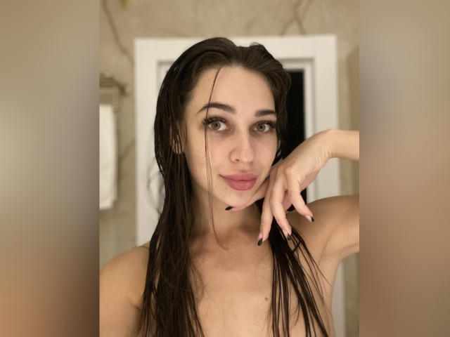 MissXPinkX, con el cabello castaño mojado, pechos pequeños y labios rosados, su mano derecha tocándose la barbilla, uñas oscuras, mirando a la cámara, en un baño..