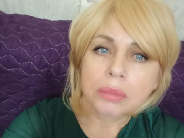 SusieNoreen has blonde hair, blue eyes, and pink lips. She's wearing a green top, looking slightly confused. Her boobs are not visible. She's against a purple quilted background.