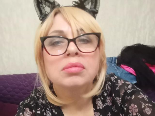 SusieNoreen with blonde hair, black cat ears, red glasses, pink lips, and a floral black shirt, showing a neutral face. Her tits are small, and she's cute.