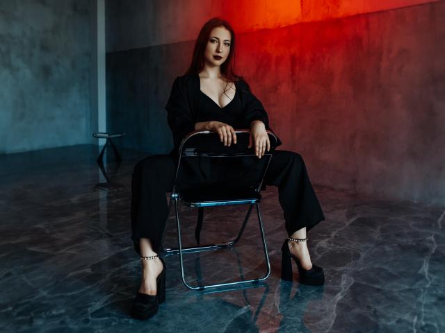FireInMySoule sits on a metal chair, wearing black pants and a deep V-neck top, with her tits slightly visible, black heels, and red lighting in the background.