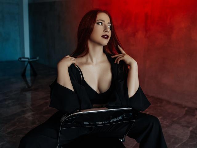 FireInMySoule has red hair, dark lipstick, and a deep cleavage in a black dress, holding the straps with hands, sitting on a shiny black chair, red light.