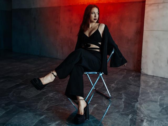 FireInMySoule sits on a chair, wearing black clothes with cutouts, showing tits, black high heels with ankle chains, and dark lipstick, in a dimly lit room.