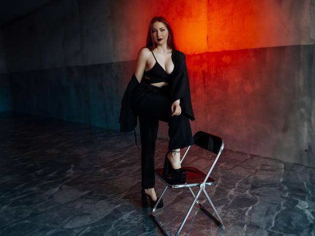 FireInMySoule with long brown hair, black bra, black pants, black high heels, black blazer off-shoulder, standing on metal chair, red and orange wall light behind.