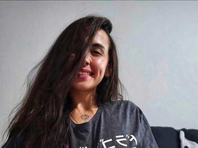 RossyNolen with long, messy black hair covering one eye, smiling, wearing a grey shirt, and a necklace with a small, visible tattoo on her chest.