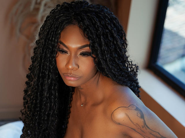 JuliethNasty has dark curly hair, brown skin, and a tattoo on her right shoulder. She's topless, showing her bare boobs and a small nipple piercing. Her eyes are closed.