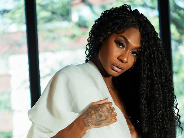 JuliethNasty with dark skin, curly black hair, and a tattoo on her right hand, wearing a white shirt, looking down with a serious expression, showing her small tits.