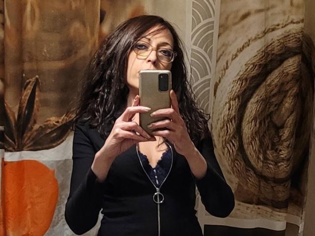 Cherazade, con i capelli castano ondulati, gli occhiali e una maglia nera con una cerniera argentata, tiene in mano un telefono e sta scattando un selfie allo specchio, mostrando i suoi piccoli seni e un reggiseno in pizzo..