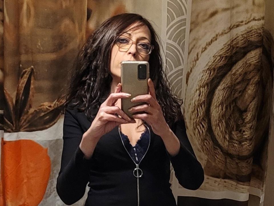Cherazade with wavy brown hair, glasses, and a black top with a silver zipper, holding a phone, taking a mirror selfie, showing her small tits and lace bra.