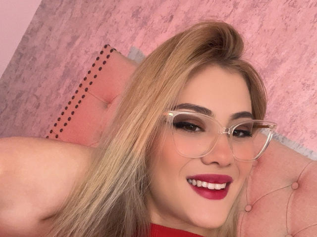 Adela Laurent, con cabello rubio, gafas de montura clara, lápiz labial rojo y una sonrisa, mostrando sus senos en una blusa roja, frente a un cabecero rosa, con borlas y adornos..
