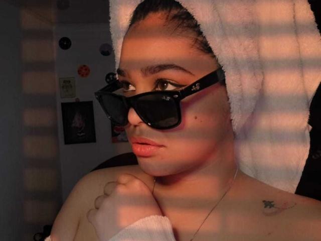 Shalyn with a white towel on her head, wearing black sunglasses, has a small tattoo on her right shoulder, and her hand rests near her chest. Soft shadows on her face.