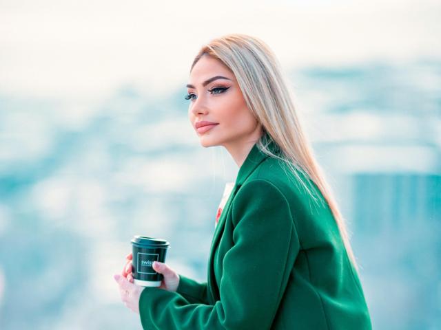 Imelda with blonde hair, green blazer, holding camera, side profile, blue eyes, winged eyeliner, subtle smile, pink lipstick, red nails, soft light, calm expression.