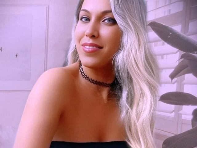 MyaHott has long, wavy blonde hair, a black choker, and is smiling with pink lipstick. She's showing her bare shoulders and a cute, sexy face.