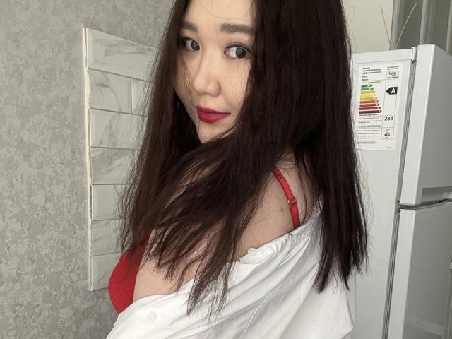DebbieLu with long dark hair, red bra, and white shirt, looking over her shoulder, showing her tits, red lipstick, and a slight smile. Refrigerator in the background.
