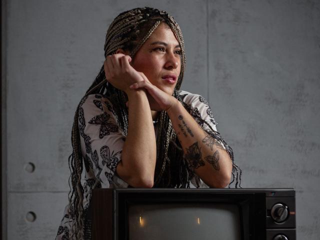 HellenK com cabelo trançado, tatuagens de borboletas nos braços, camisa branca com borboleta, apoiada em uma TV antiga, mãos sob o queixo, olhando para o lado, batom rosa, parede de concreto ao fundo..