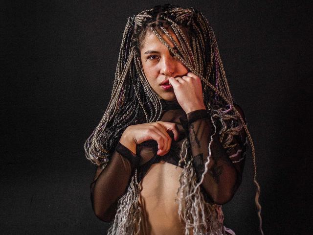 HellenK met lang gevlochten haar, gekleed in een doorschijnend zwart topje, raakt haar lippen aan met haar rechterhand, waarbij haar kleine borsten zichtbaar worden. Donkere achtergrond, gefocust op haar bovenlichaam..