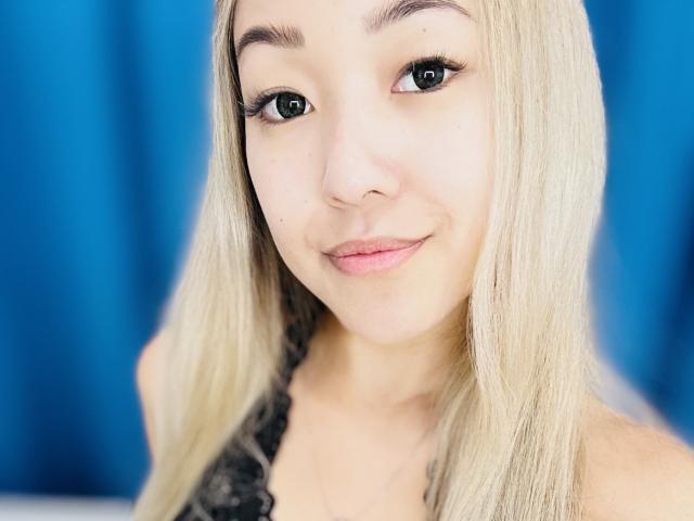 RenyLime has light blonde hair, green eyes, and a slight smile, wearing a black lace top, with a soft, cute face and subtle makeup.