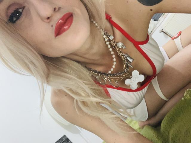 MissCherryOnTop, avec ses cheveux blonds, son rouge à lèvres rouge et son collier de perles, portant un haut blanc et rouge, montrant ses seins et sa culotte blanche, est agenouillée sur une serviette verte..