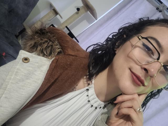 Gabriela Moore, con cabello negro rizado, gafas y una sonrisa discreta, vestida con un vestido blanco y un collar de cuentas negras, un abrigo marrón, y un gato con una cabeza marrón y peluda..
