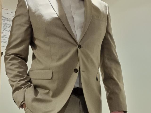RicRockhard in a beige suit jacket, white shirt, and beige pants, with hands in pockets, showing a clean-chest and dark buttons on the jacket.