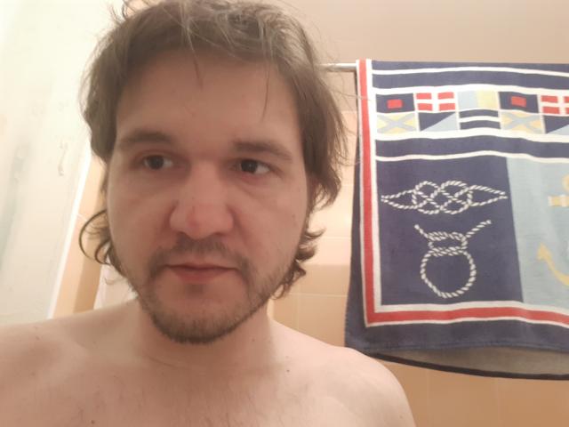 NectarineFad has a chubby face, brown wavy hair, light beard, and is shirtless with visible chest hair. A blue towel with white and red patterns hangs behind.