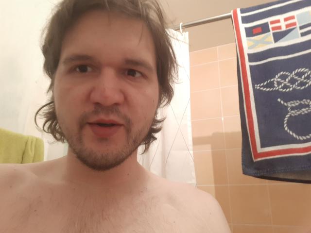 NectarineFad has a chubby face, brown wavy hair, and a beard, standing shirtless with a slight smirk, showing a hairy chest, in a bathroom.