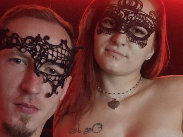 LesLioups with a black lace mask, red-haired, topless, showing tits, and a heart necklace, standing next to LesLioups with a similar mask, slightly smiling.