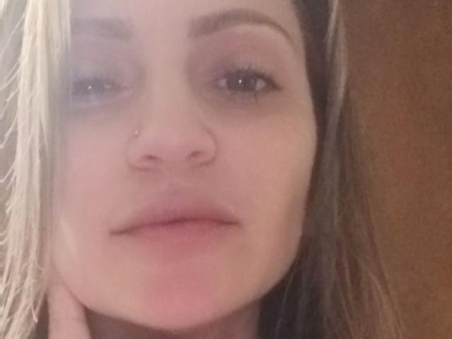 AliaAnita's face with blonde hair, slight smile, light pink nose piercing, subtle pink lips, and brown eyes. Her hand rests near her chin. Soft, warm lighting.