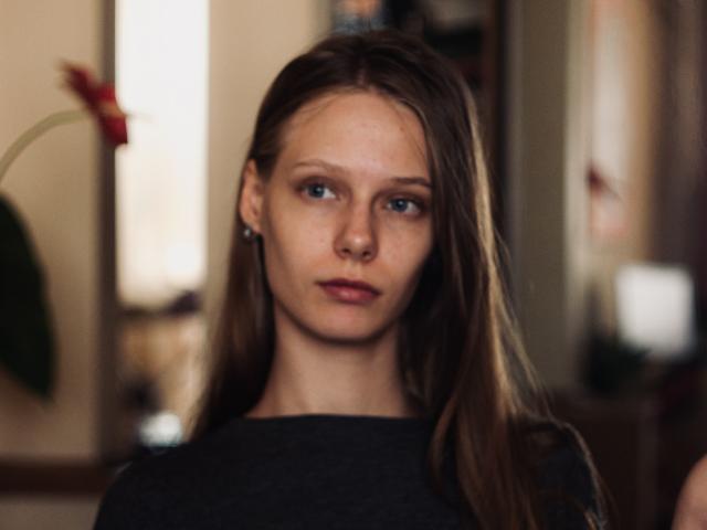 Lallilillo a de longs cheveux bruns, des yeux bleus et une expression sérieuse. Elle porte une chemise sombre, et ses petites seins sont légèrement visibles. Elle se trouve dans un environnement intérieur flou..