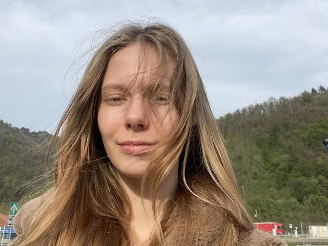 Lallilillo has long, wind-swept blonde hair covering her eyes, a slight pout, and a cute face with light freckles, wearing a brown fuzzy coat, in a cloudy outdoor setting.