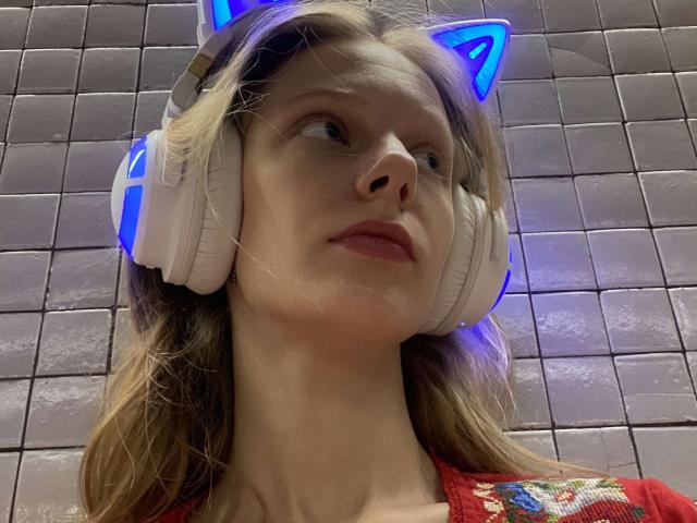 Lallilillo with blonde hair, blue cat ears, and white headphones, looking up with a slight smirk, wearing a red shirt with colorful embroidery, in a tiled room.