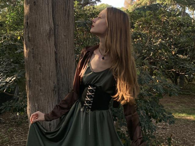 Lallilillo, avec de longs cheveux bruns, les yeux fermés, la tête penchée en arrière, portant une robe verte avec un corsage noir à lacets, se tient près d'un arbre, la lumière du soleil sur son visage, tenant sa robe..
